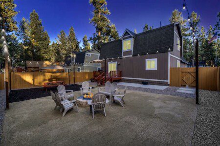 Cozy outdoor fire pit area with wooden Adirondack chairs in the backyard of a modern vacation rental home surrounded by tall pine trees.