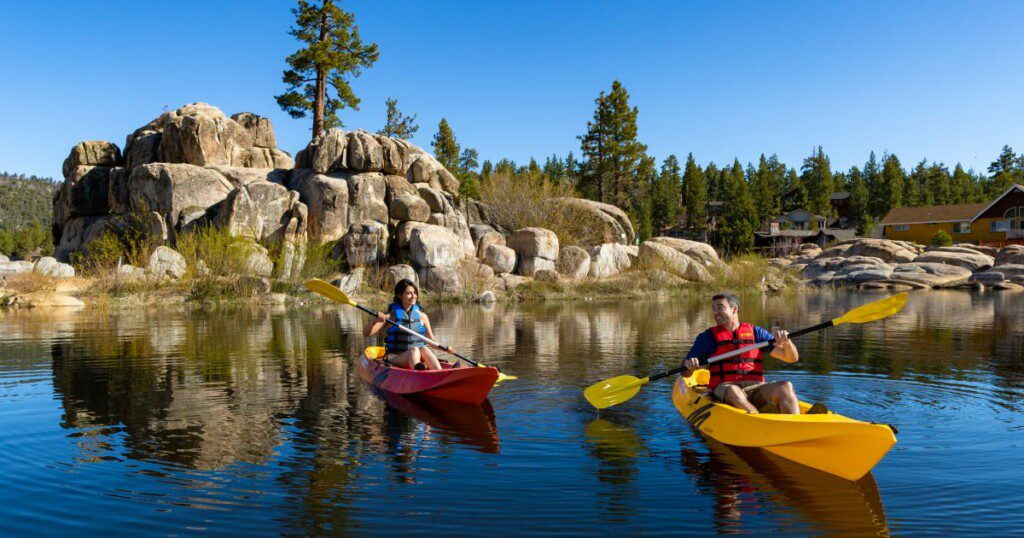 "Scenic view of Big Bear Lake with kayakers paddling in the calm waters surrounded by lush, green forests and mountains in the background, highlighting the beauty of eco-friendly travel in Big Bear, CA."