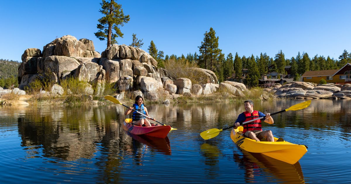 "Scenic view of Big Bear Lake with kayakers paddling in the calm waters surrounded by lush, green forests and mountains in the background, highlighting the beauty of eco-friendly travel in Big Bear, CA."
