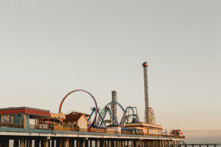 Galveston Island Historic Pleasure Pier amusement park with Iron Shark roller coaster, Galaxy Wheel Ferris wheel, and carnival rides over the Gulf of America at sunset